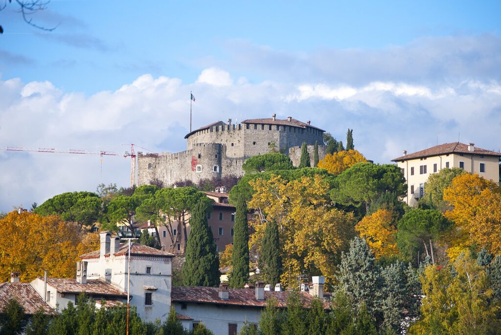 Il castello di gorizia in prima piano è circondato da una ricca vegetazione. Il cielo è azzuro con qualche nuvola.