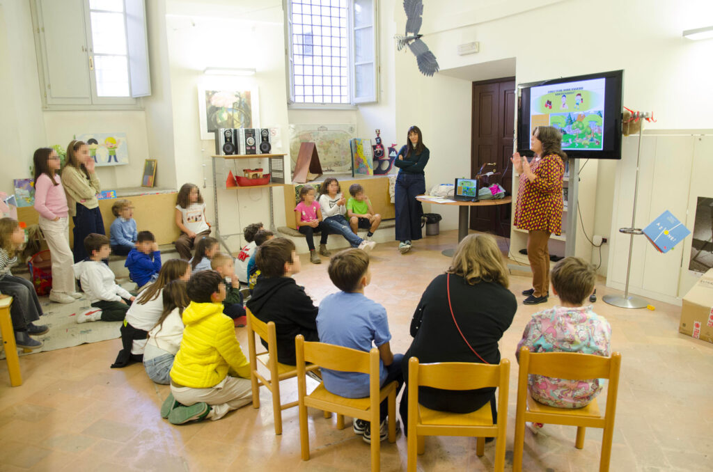 Bambini di spalle ascoltano una donna adulta che illustra delle immagini in TV