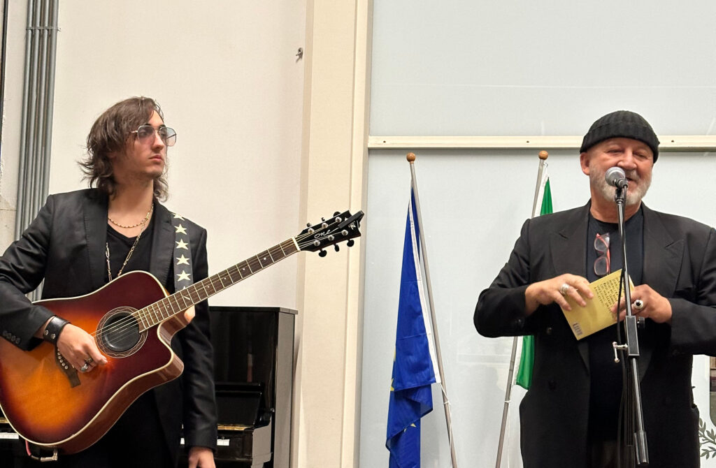Un musicista con chitarra acustica e un uomo al microfono aprono l’evento della mostra “L’eredità della vita” a Marcallo con Casone.