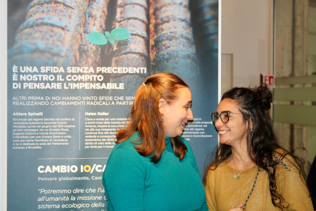 Due visitatrici sorridono davanti a un pannello della mostra “L’eredità della vita” con il testo dedicato alle sfide e ai cambiamenti globali.