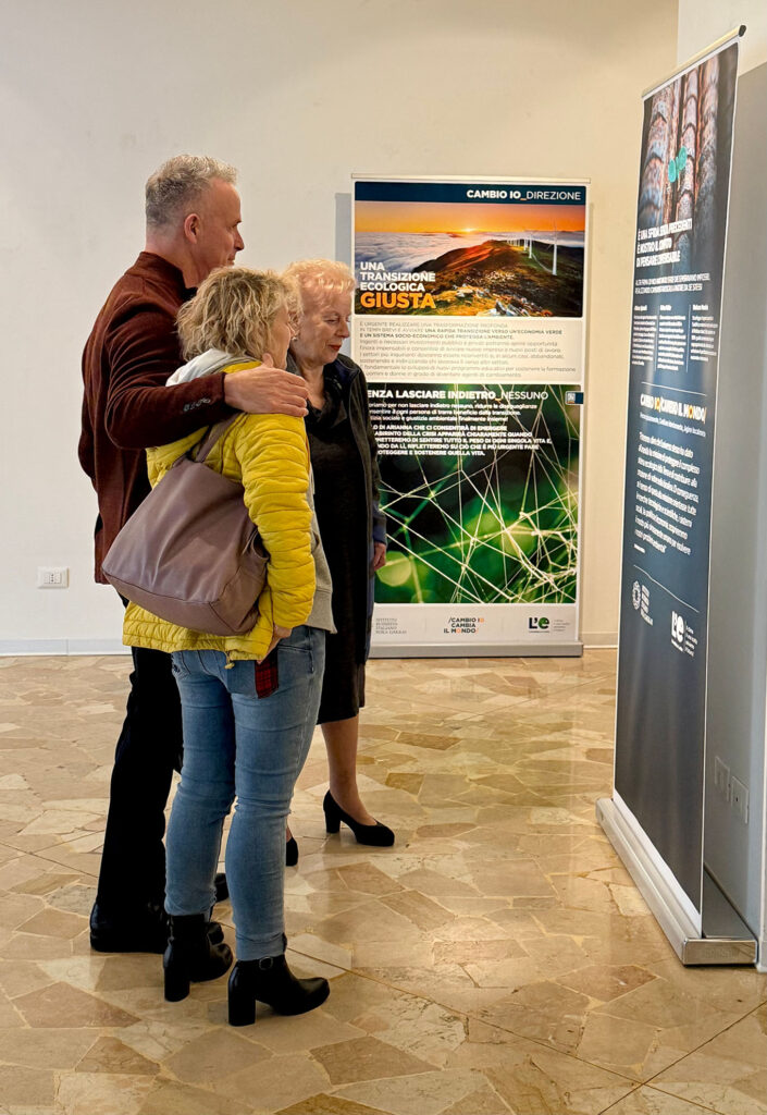 Tre persone osservano insieme i contenuti espositivi della mostra “L’eredità della vita” allestita nella Sala Tullio Crali di Gorizia.