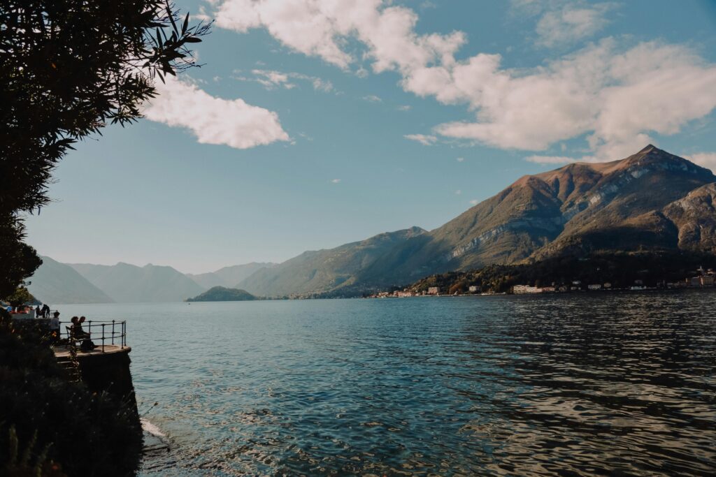 Immagine del paesaggio lombardo. Il primo piano il lago di Come con dietro delle montagne. Il cielo è limpido con qualche nuvola.