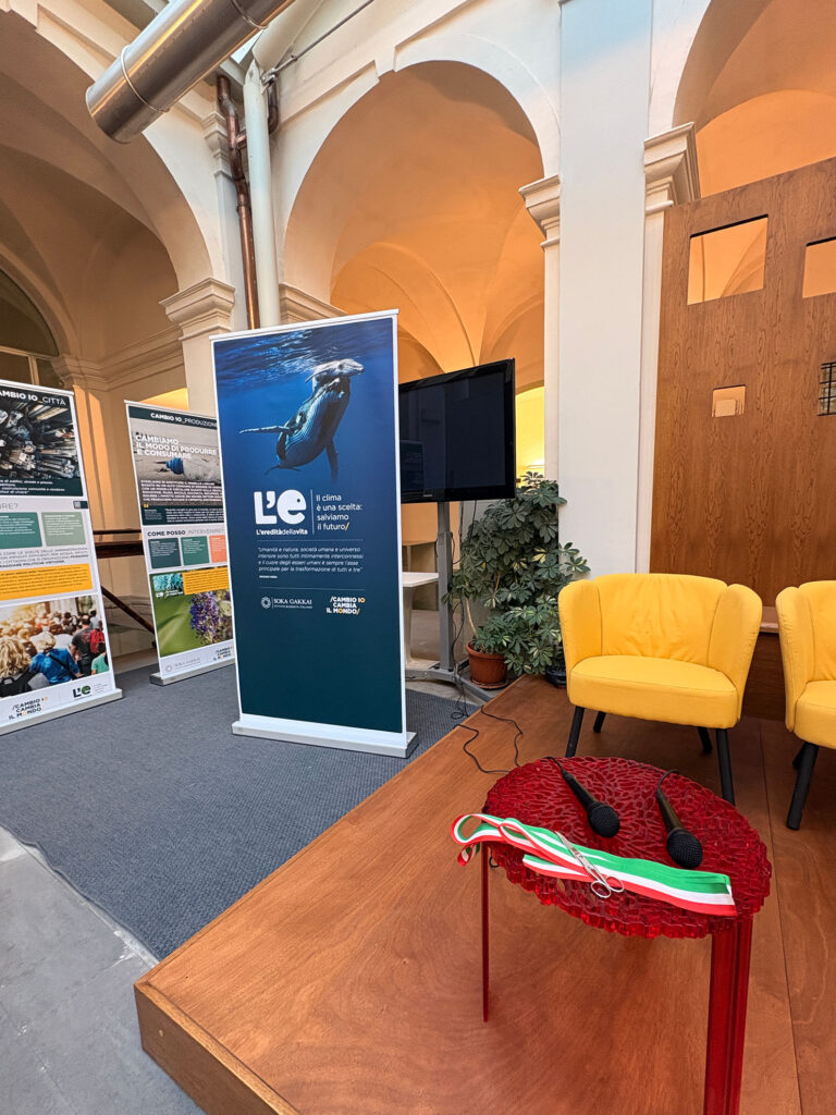 Allestimento per l'inaugurazione della mostra in un edificio con architettura a volte. In primo piano un piccolo tavolo rosso con sopra due microfoni, un paio di forbici e un nastro tricolore italiano, suggerendo una cerimonia d’inaugurazione. Sullo sfondo due poltrone gialle, uno schermo nero e pannelli informativi dedicati a temi ambientali e climatici con il logo “L’E — L’eredità della vita"