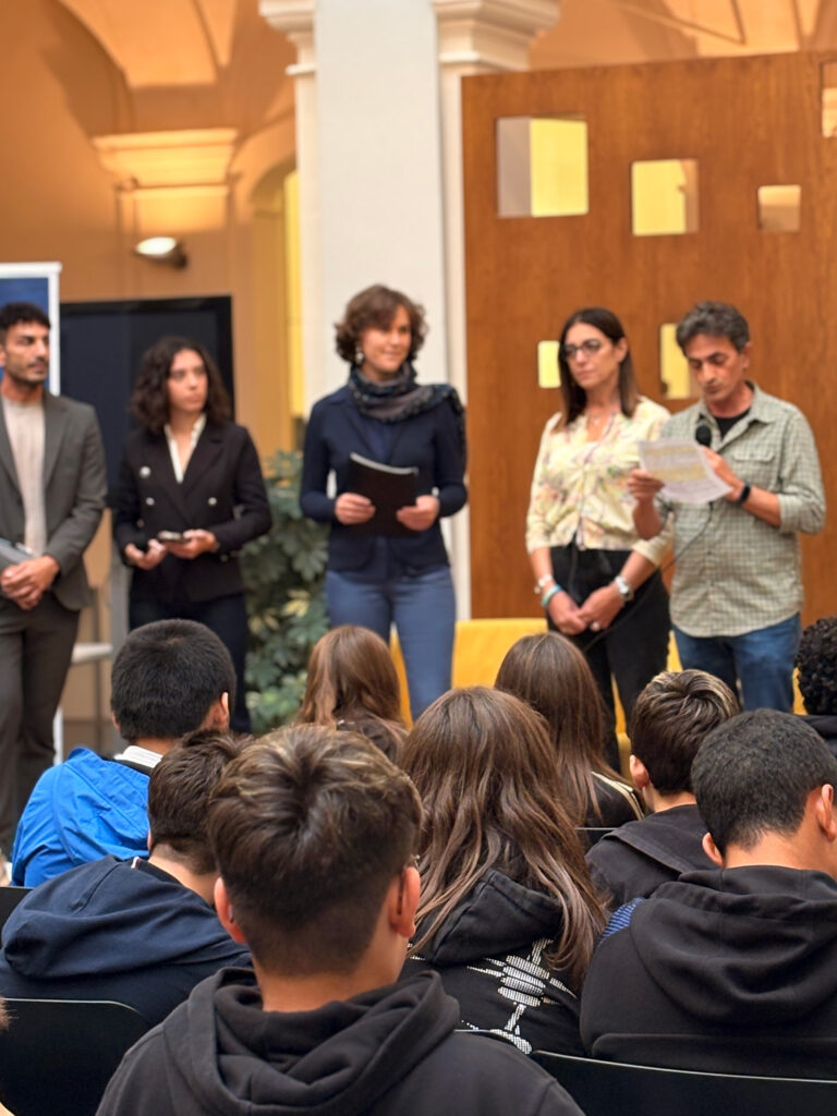 Un gruppo di persone in piedi davanti a un pubblico durante un evento in un edificio con architettura a volte. Alcuni dei relatori tengono fogli o tablet, mentre una persona parla al microfono. In primo piano si vedono diversi ragazzi seduti, di spalle, che assistono alla presentazione.