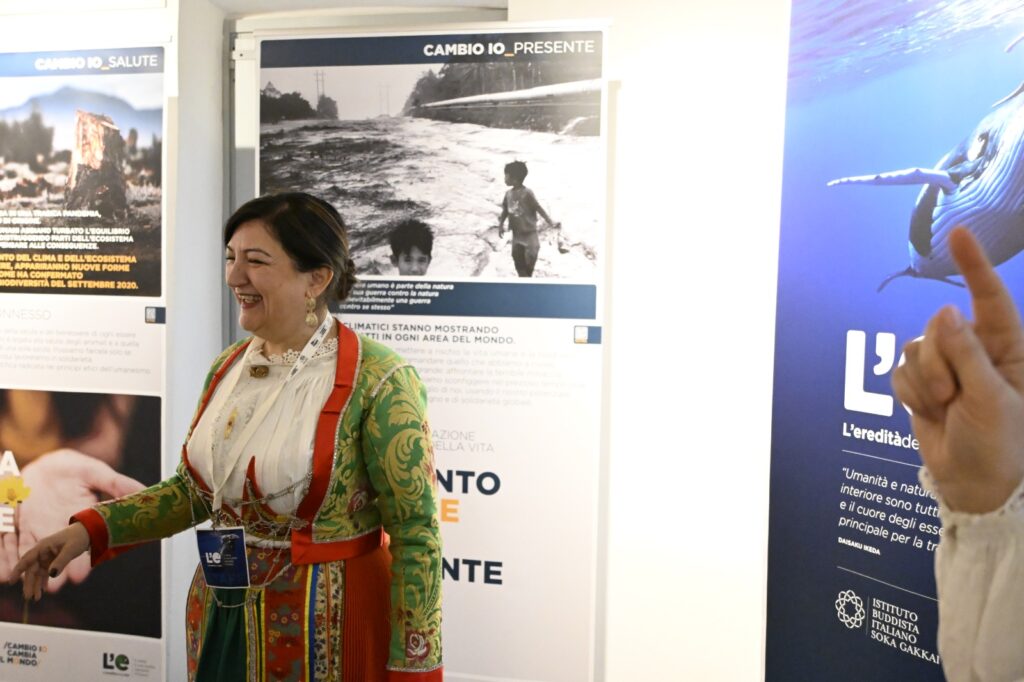 Donna in abito tradizionale sardo visita la mostra “L’eredità della vita”, davanti a pannelli informativi sul clima e l’ambiente.