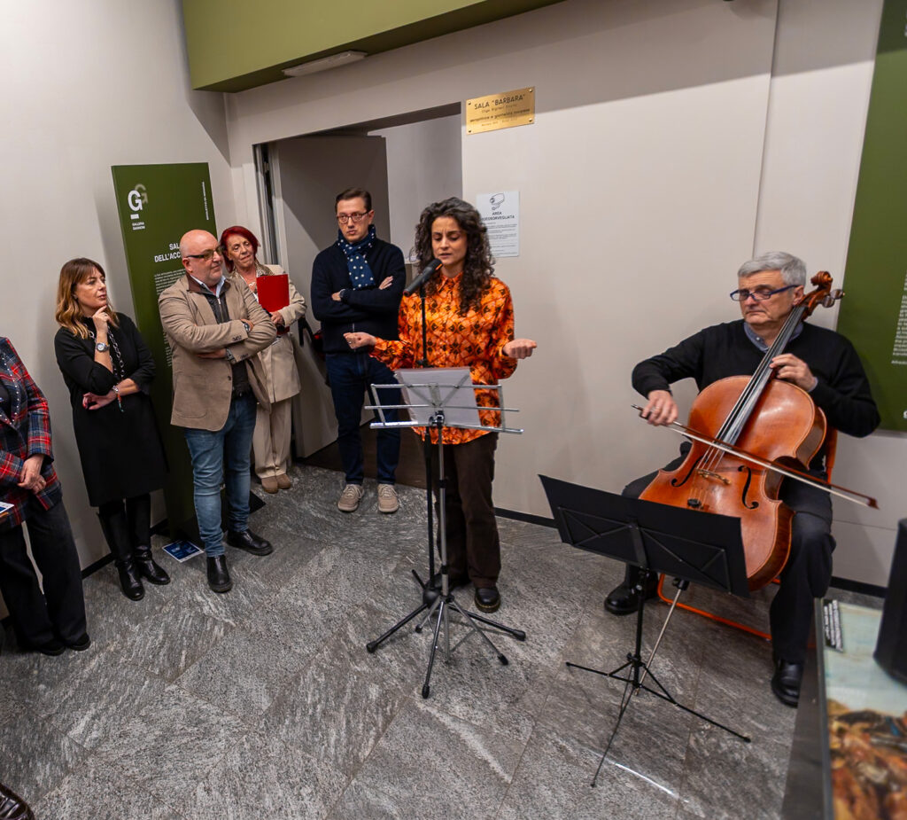 Persona legge a un leggio durante l’inaugurazione della mostra mentre un musicista suona il violoncello; alcune persone ascoltano accanto all’ingresso della sala.