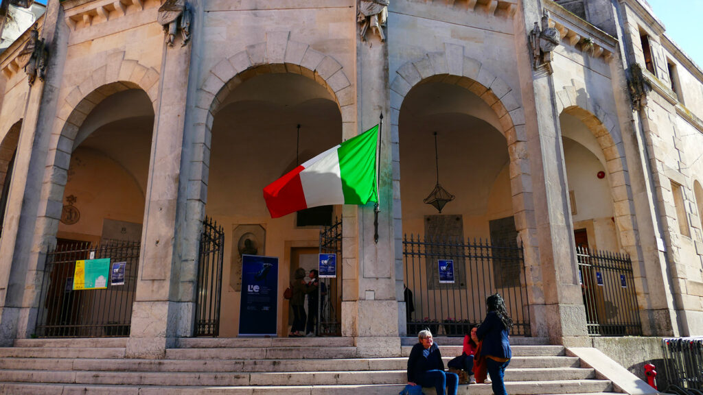 Facciata della Sala della Gran Guardia con portico e bandiera italiana, ingresso della mostra con locandine esposte.