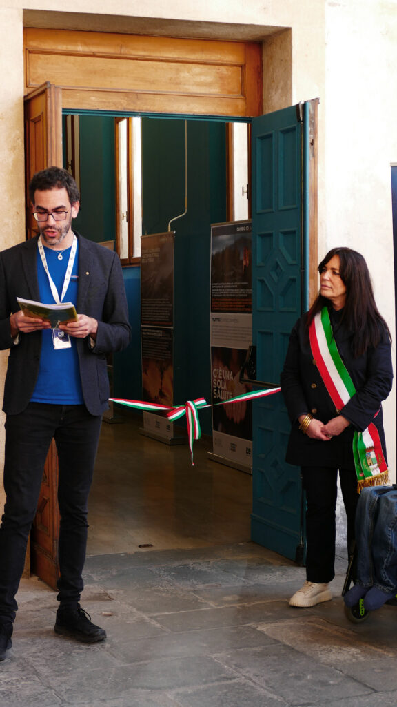 Persona con badge legge un intervento accanto a una rappresentante istituzionale con fascia tricolore, davanti all’ingresso della mostra.
