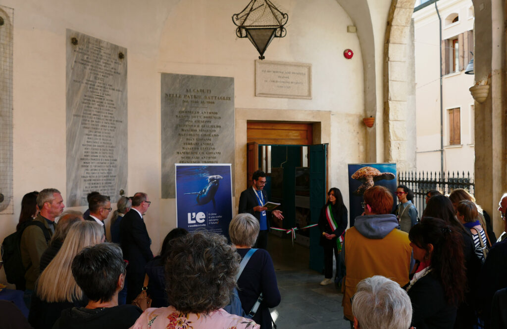 Pubblico riunito ascolta un intervento all’ingresso della mostra, con pannelli espositivi e porta aperta sullo sfondo.