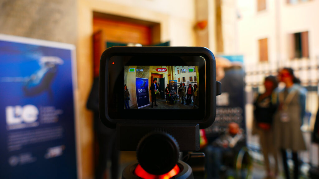 Telecamera in primo piano che riprende un momento della presentazione della mostra con pubblico sullo sfondo.