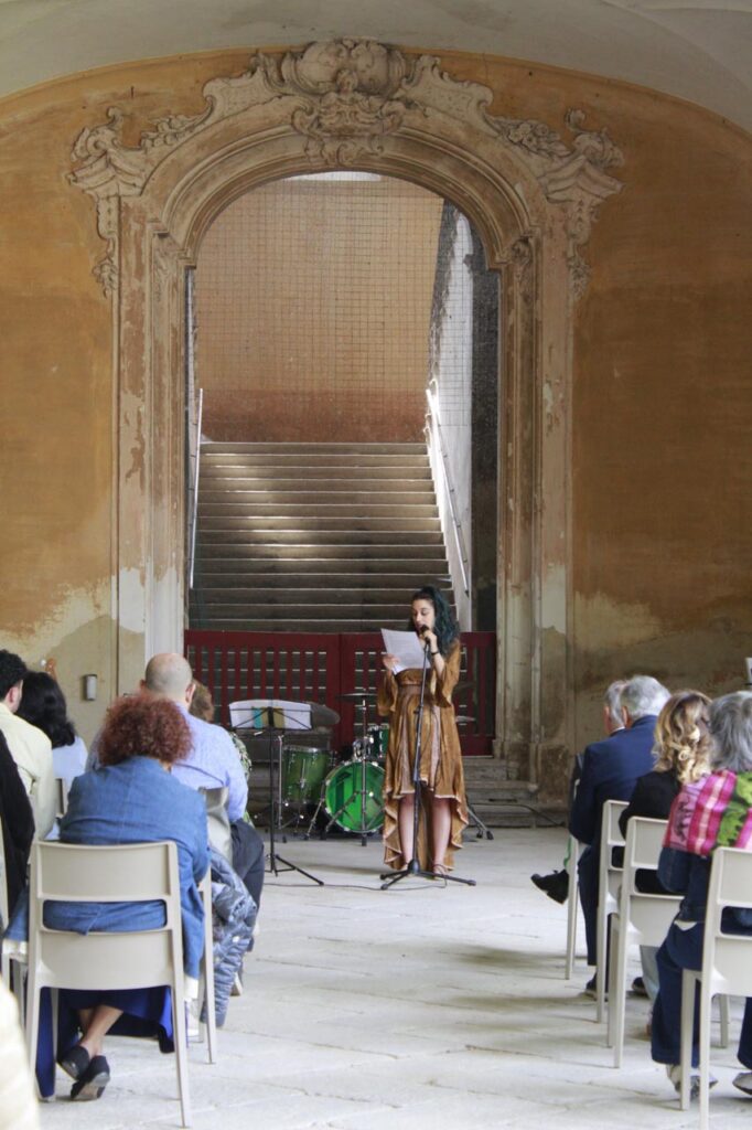 Persona legge un testo al microfono davanti al pubblico durante l’inaugurazione.